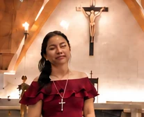 Florane in a red dress singing with eyes closed in a chapel, a crucifix behind her